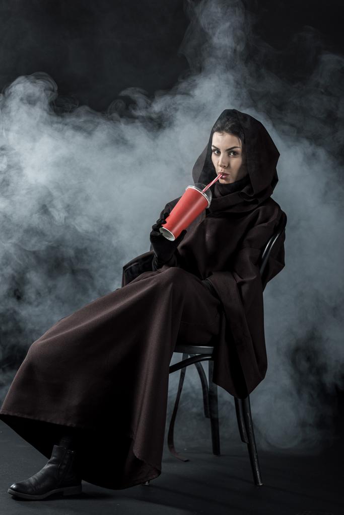 vrouw in de dood kostuum zitten op stoel en drinken drank op zwart - Foto, afbeelding