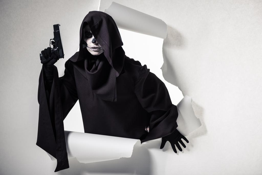 женщина в костюме смерти держит пистолет и вылезает из дыры в бумаге
 - Фото, изображение