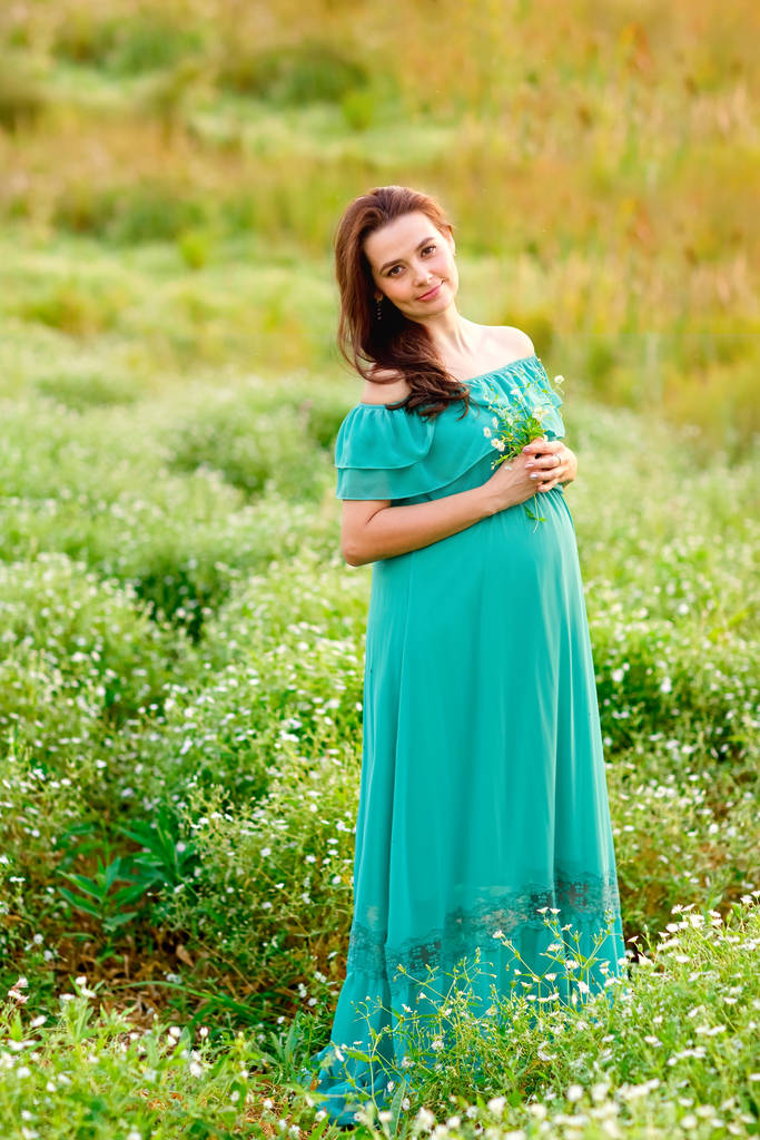 schöne schwangere Frau in grünem Kleid steht im Weidengarten und blickt an einem Sommertag in die Kamera - Foto, Bild