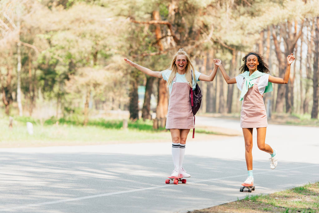 full length view of two smiling multicultural friends holding hands while skateboarding on road - Photo, Image