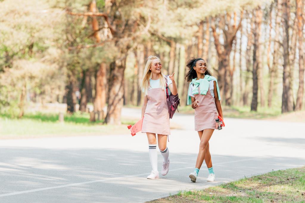 Ganzkörperansicht von zwei lächelnden multikulturellen Freunden, die Penny-Bretter in der Hand halten und auf der Straße gehen - Foto, Bild