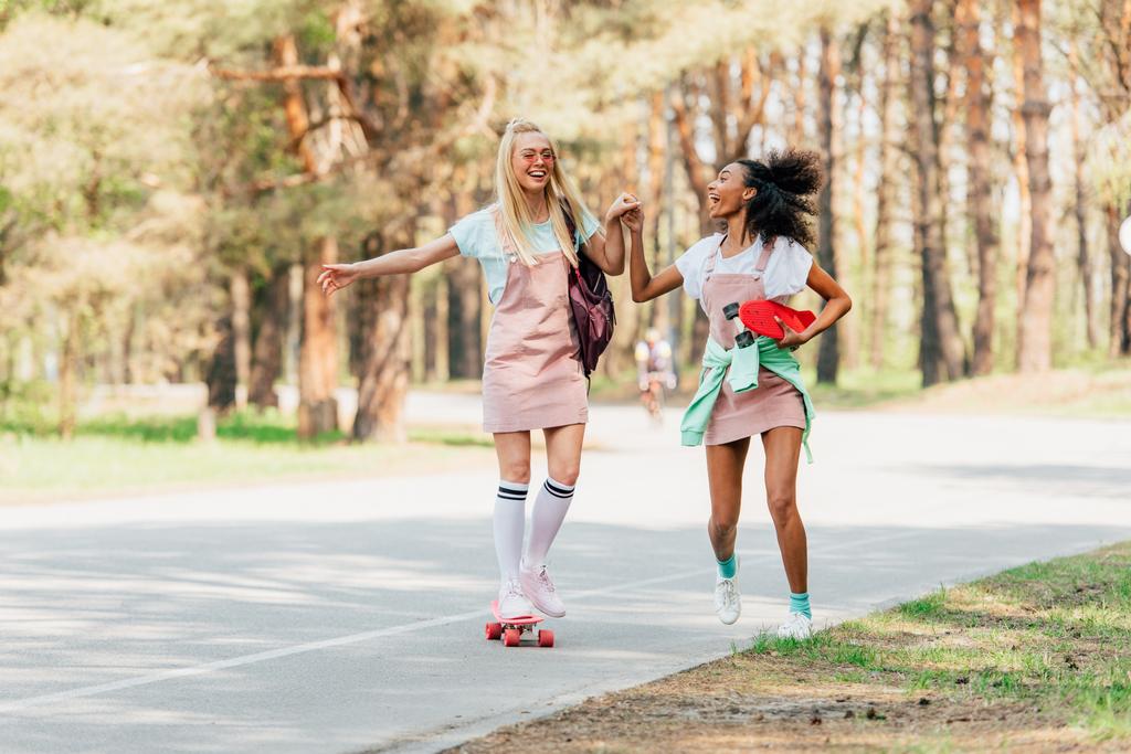 vista completa de dos amigos multiculturales sonrientes tomados de la mano mientras patinan en la carretera
 - Foto, imagen