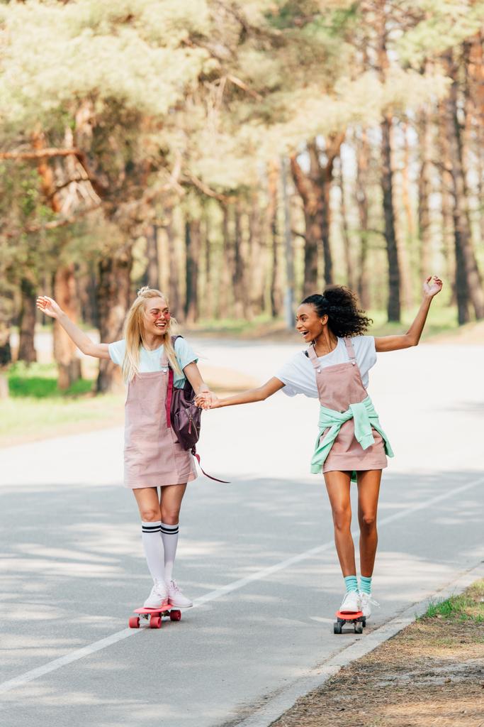 visão de comprimento total de dois amigos multiculturais sorridentes de mãos dadas enquanto skate na estrada
 - Foto, Imagem