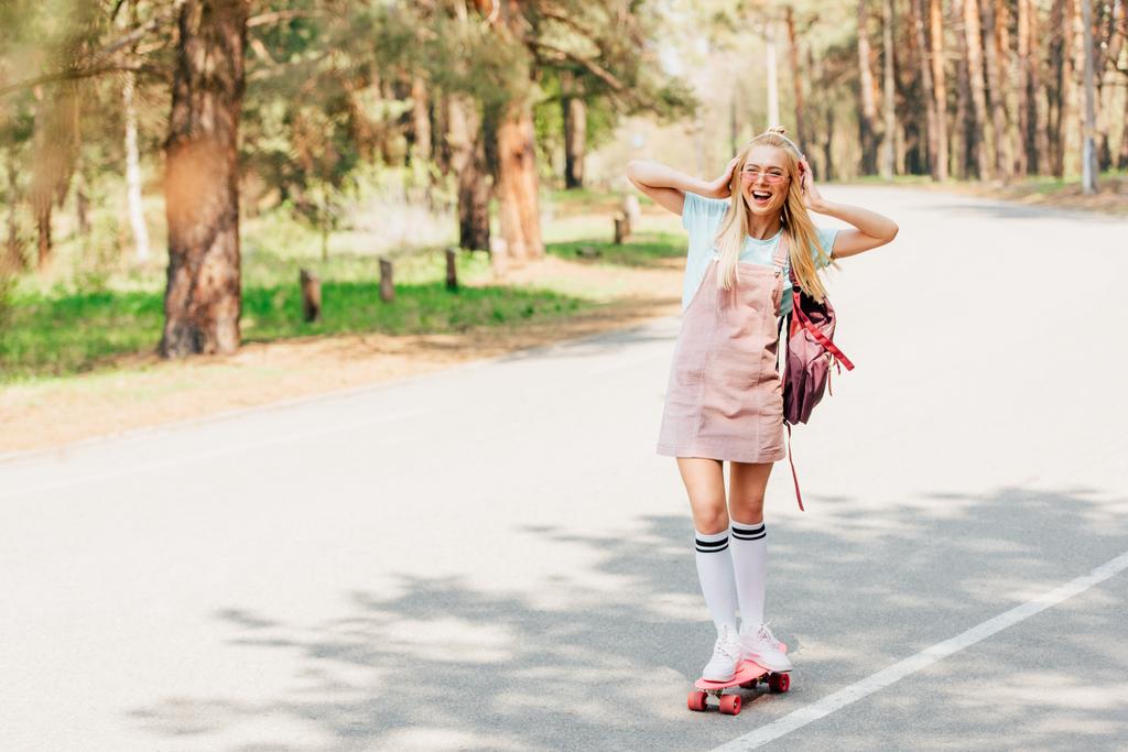 Ansicht des aufgeregten blonden Mädchens beim Skateboarden und Musikhören über Kopfhörer in voller Länge - Foto, Bild