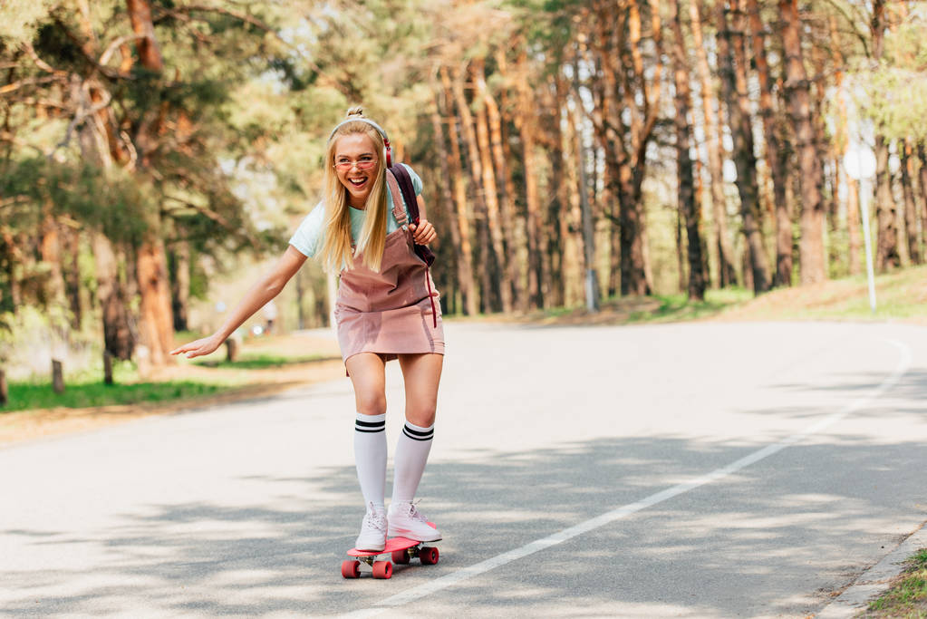 full length view of excited blonde girl skateboarding and listening music in headphones - Photo, Image