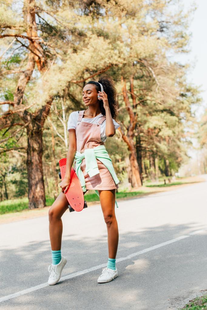 visão de comprimento total da menina americana africana em fones de ouvido de pé na estrada e segurando penny board
 - Foto, Imagem