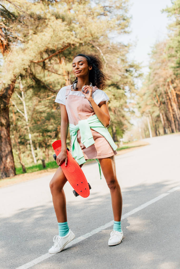 visão de comprimento total da menina americana africana em fones de ouvido de pé na estrada e segurando penny board
 - Foto, Imagem