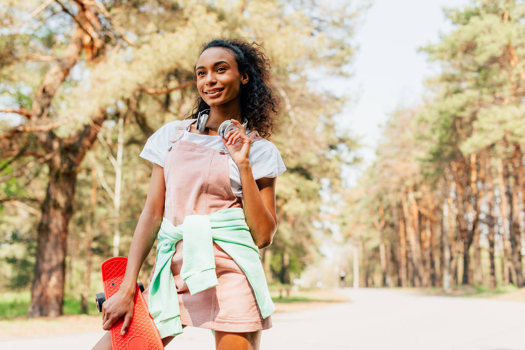 african american girl in headphones standing on road and holding penny board - Photo, Image