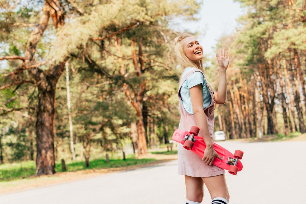 возбужденная блондинка, держащая доску с пенни и машущая рукой на дороге
 - Фото, изображение