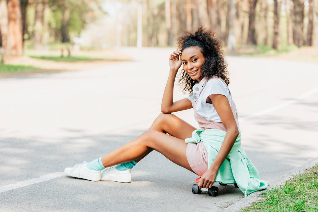 道路上のペニーボードに座ってヘッドフォンを持つ笑顔のアフリカ系アメリカ人の女の子 - 写真・画像