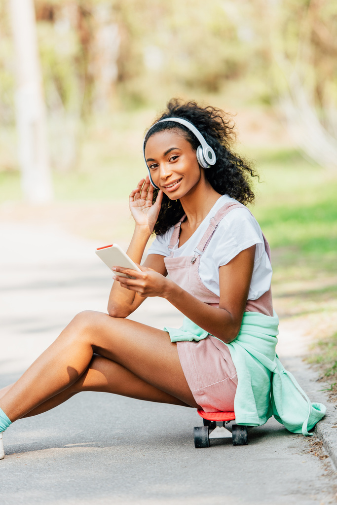 sorrindo menina afro-americana ouvindo música em fones de ouvido e usando smartphone enquanto sentado em placa de centavo
 - Foto, Imagem