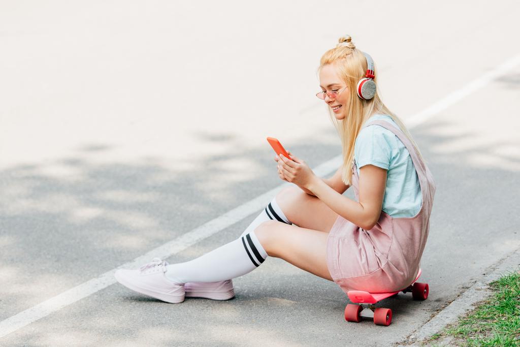 道路上のペニーボードに座っている間、ヘッドフォンで音楽を聴いて笑顔のブロンドの女の子 - 写真・画像