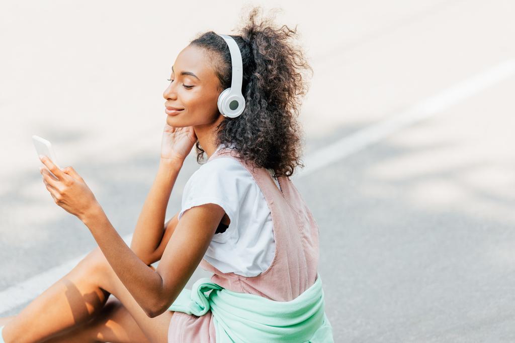 smiling african american girl listening music in headphones and using smartphone on road - Photo, Image