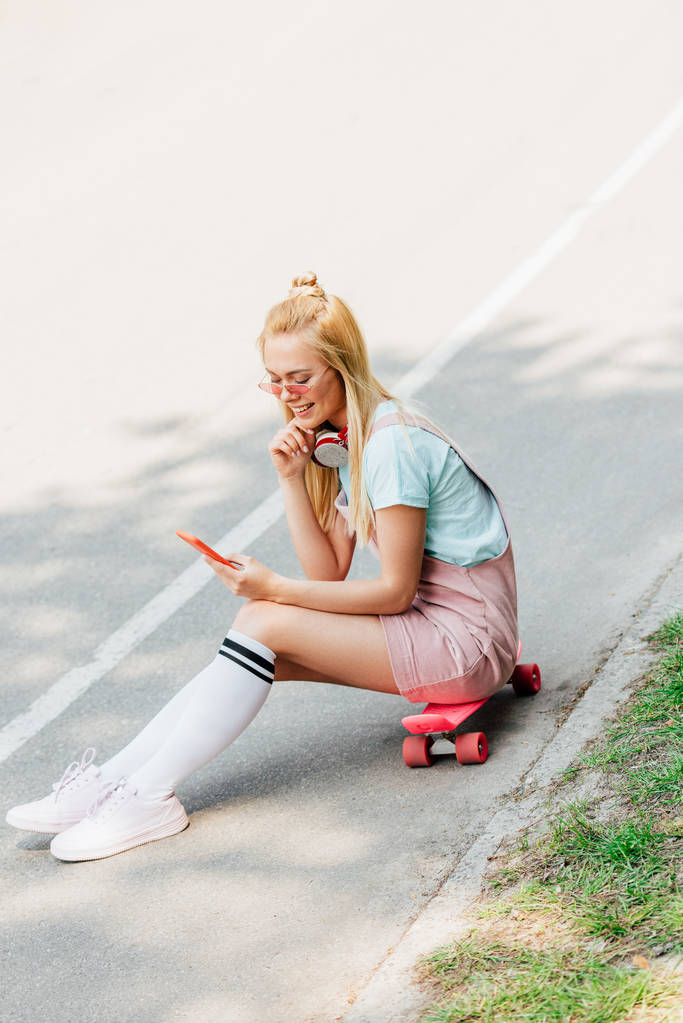 sorridente ragazza bionda ascoltare musica in cuffia mentre seduto su penny board sulla strada
 - Foto, immagini