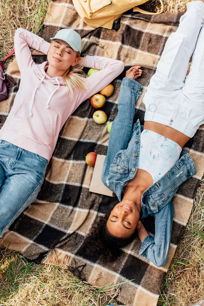 vista superior de dos chicas multiétnicas sonrientes acostadas en manta a cuadros con manzanas
 - Foto, Imagen