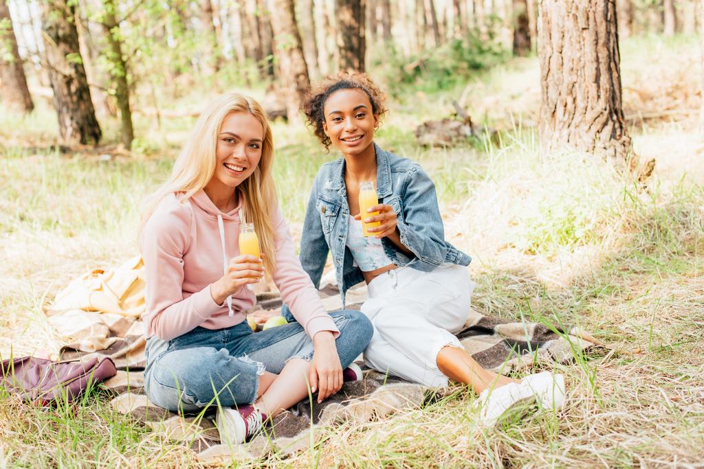  дві багатоетнічні дівчата сидять на плетеній ковдрі і тримають пляшки апельсинового соку
 - Фото, зображення