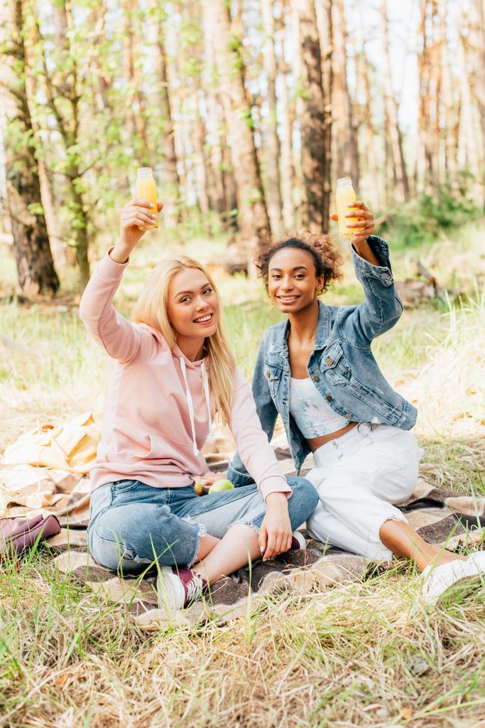  dos chicas multiétnicas sentadas en manta a cuadros y criando botellas de jugo de naranja
 - Foto, imagen