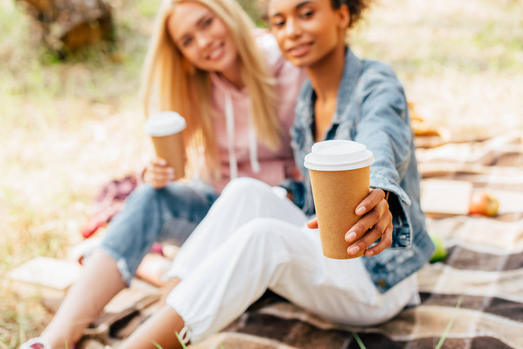 vista recortada de dos amigos multiétnicos sentados en manta a cuadros y sosteniendo tazas de papel de café
 - Foto, Imagen