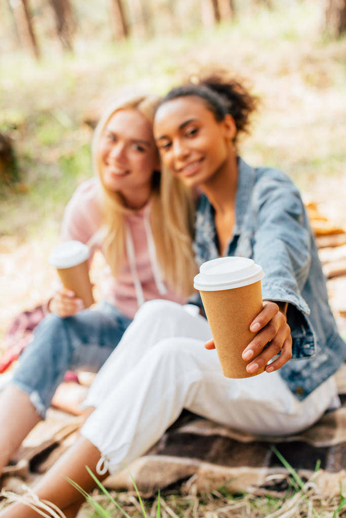 selective focus of two multiethnic friends sitting on plaid blanket and holding paper cups of coffee - Photo, Image