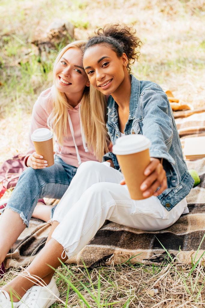  deux amis multiethniques assis sur une couverture à carreaux et tenant des tasses de café en papier
 - Photo, image