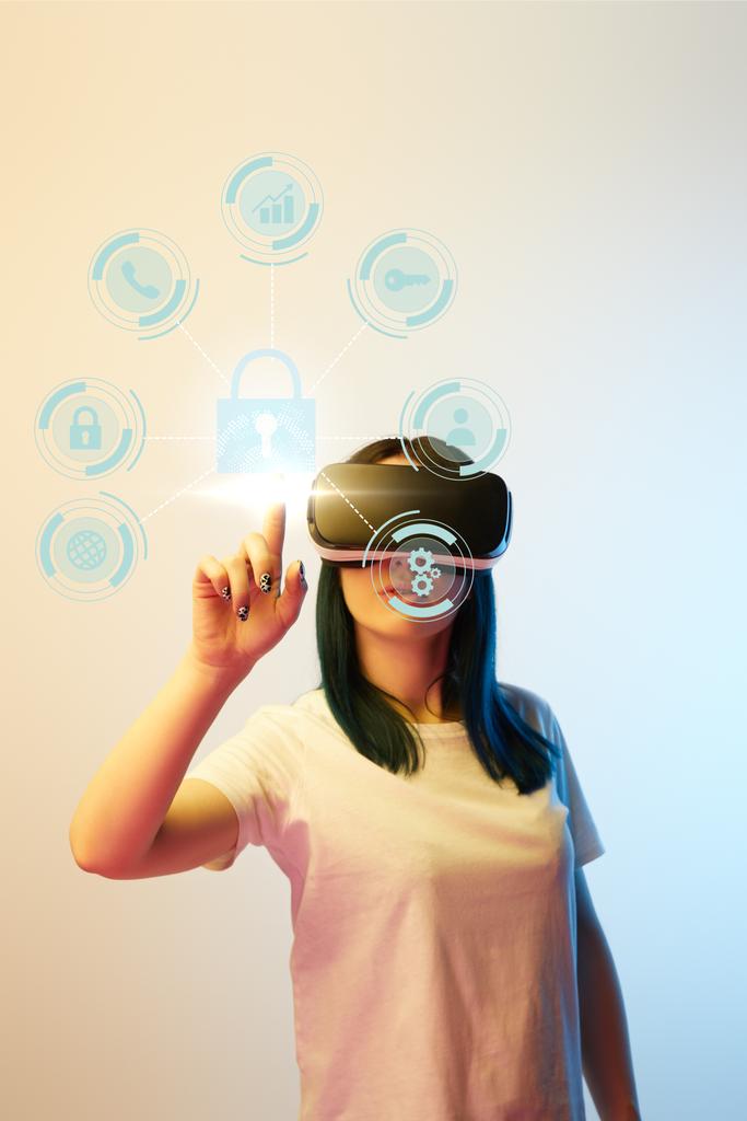 mladá žena ve virtuální realitě sluchátka ukazuje prstem na internetové bezpečnostní ikony na béžové a modré pozadí - Fotografie, Obrázek