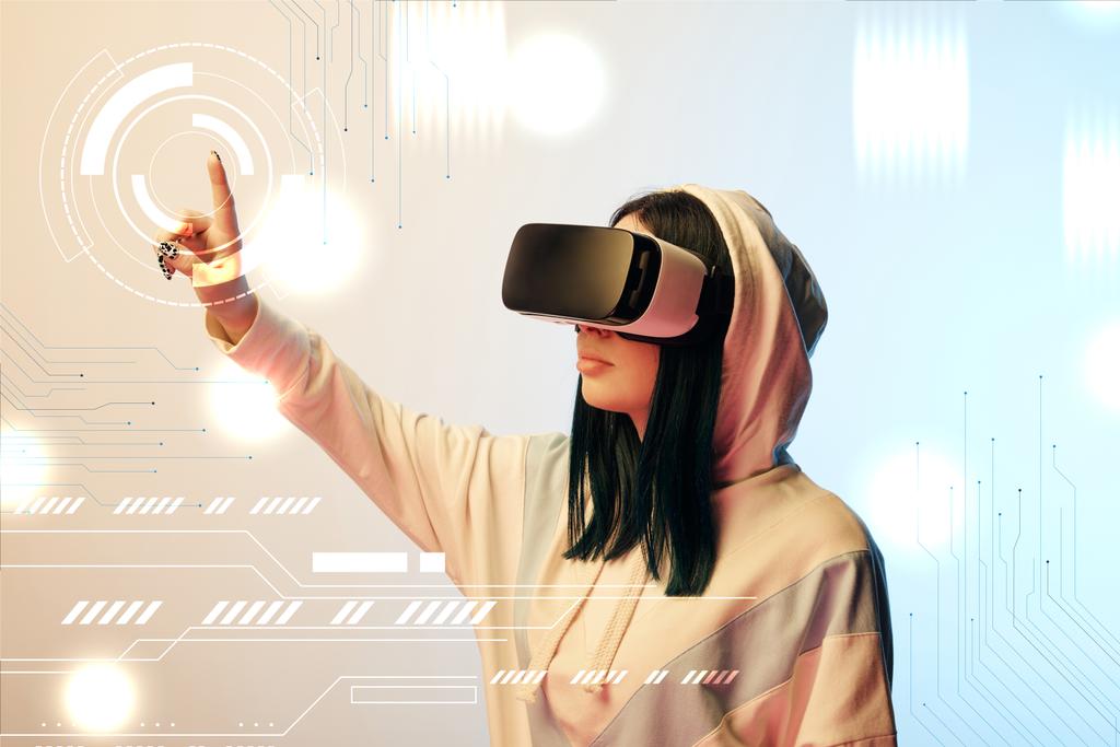 young woman in virtual reality headset pointing with finger at glowing cyber illustration on beige and blue background - Photo, Image