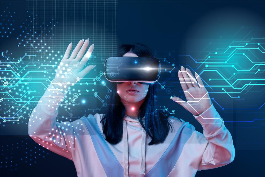 mladá žena ve virtuální realitě headset gestikulace mezi zářící kybernetické ilustrace na tmavém pozadí - Fotografie, Obrázek