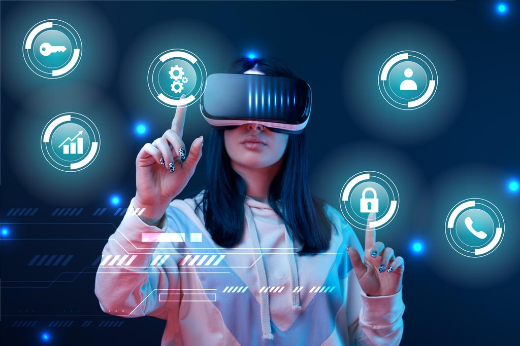 mujer joven en auriculares de realidad virtual apuntando con los dedos a los iconos cibernéticos brillantes sobre fondo oscuro
 - Foto, imagen