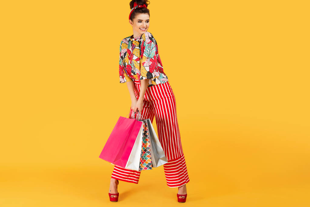 Úplná lenhts fotografie mladé šťastné kavkazské ženy v příležitostných barevných šatech držící tašky a nakupování přes oranžové pozadí. Koncepce nakupování a prodeje. - Fotografie, Obrázek
