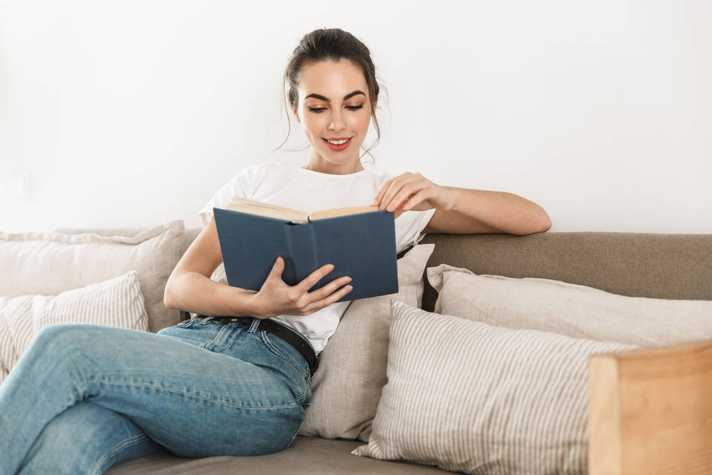 schöne glückliche junge Studentin sitzt drinnen und liest Buch auf dem Sofa. - Foto, Bild