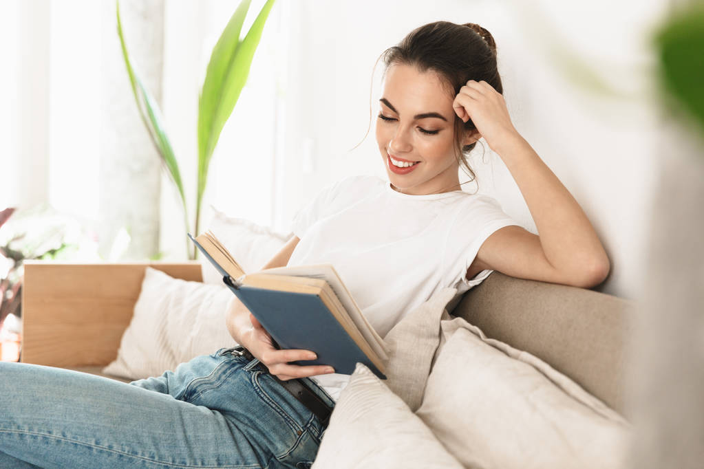 Piękny szczęśliwy młody student dziewczyna siedzący w pomieszczeniu czytanie książka na sofa. - Zdjęcie, obraz