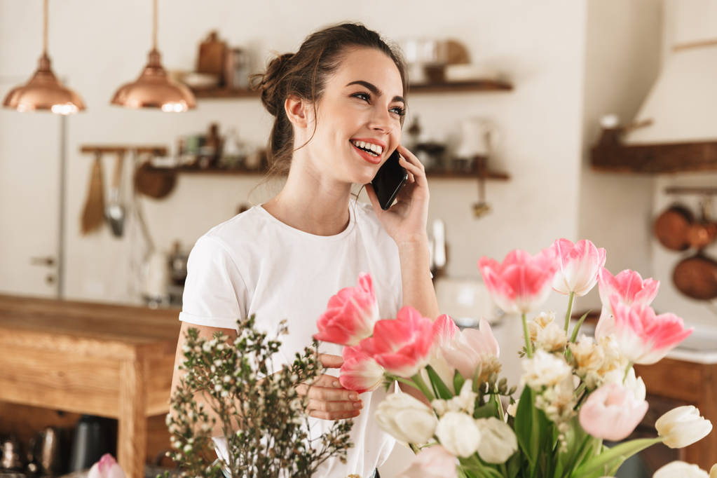 Hermosa joven estudiante feliz posando en el interior cerca de flores hablando por teléfono móvil
. - Foto, imagen