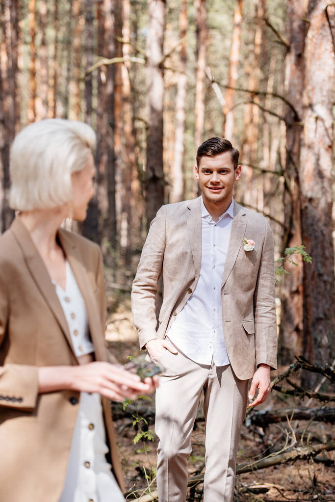 χαμογελαστός γαμπρός στέκεται με το χέρι στην τσέπη και κοιτάζοντας τη νύφη στο δάσος - Φωτογραφία, εικόνα