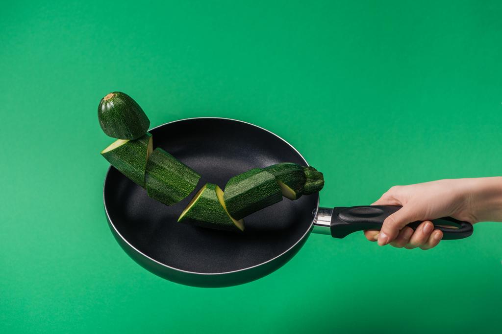 Partial view of woman holding frying pan with fresh green sliced zucchini on green background - Photo, Image