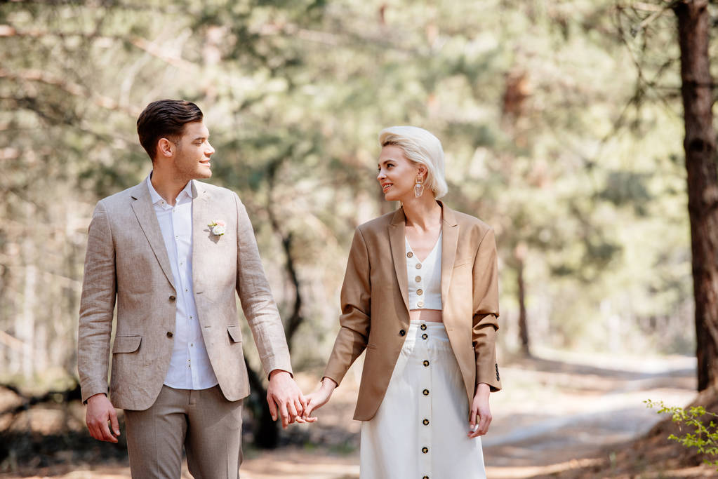 happy stylish couple holding hands and looking at each other in forest - Photo, Image