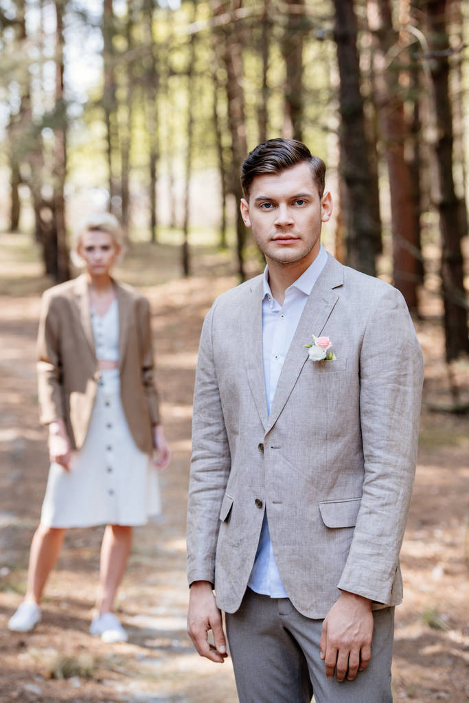 elegante sposo con boutonniere su giacca e sposo in piedi nella foresta e guardando la fotocamera
 - Foto, immagini