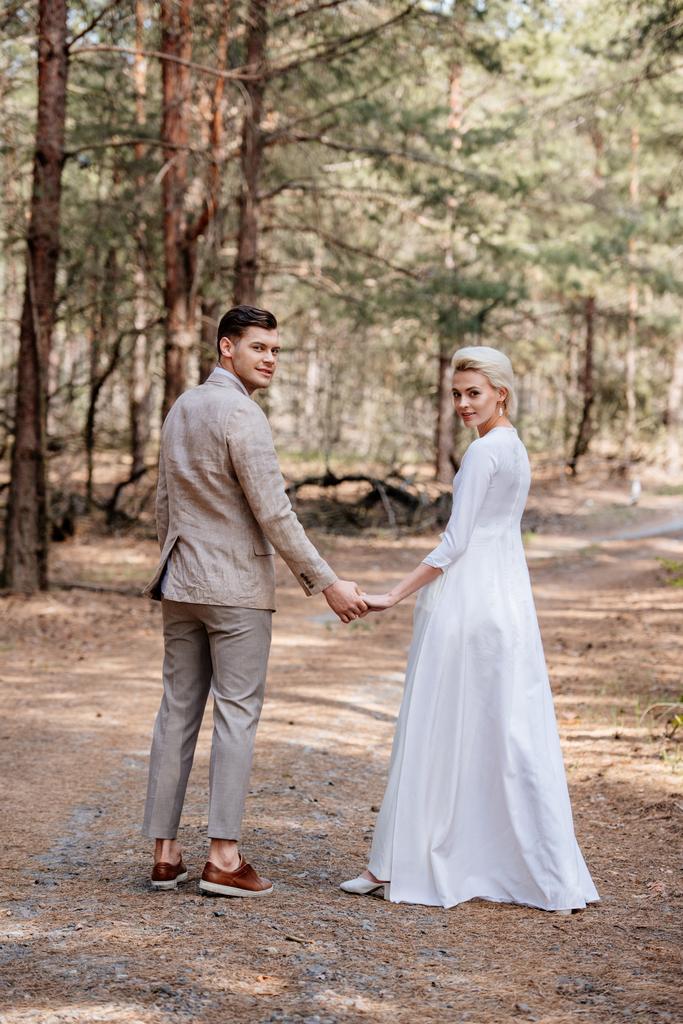 täyspitkä näkymä hymyilevä juuri aviopari kädestä metsässä
 - Valokuva, kuva