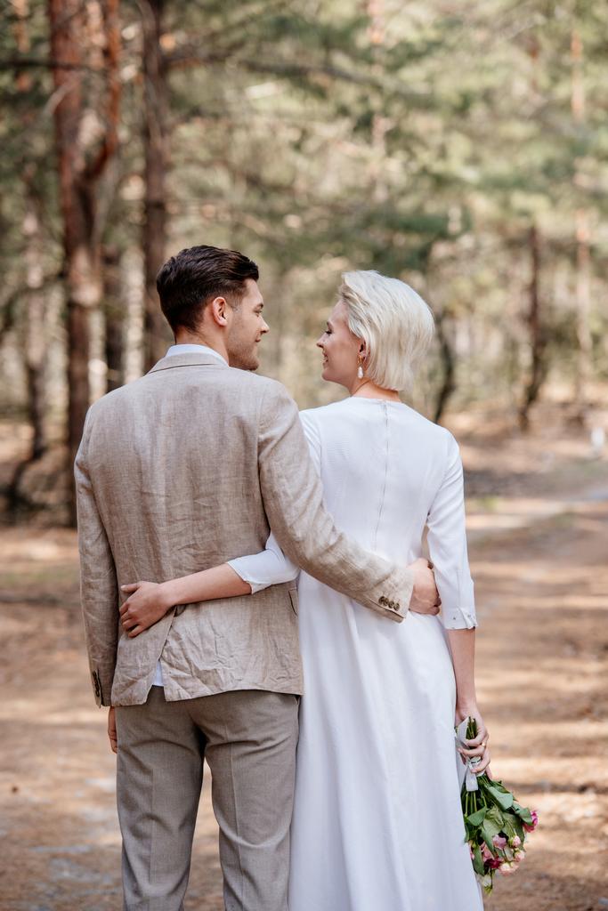 back view of just married couple embracing and looking at each other in forest - Photo, Image