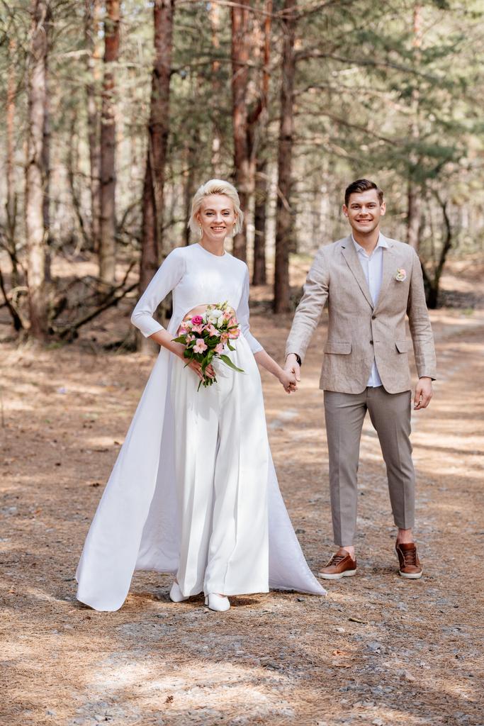 full length view of smiling just married couple holding hands in forest - Photo, Image