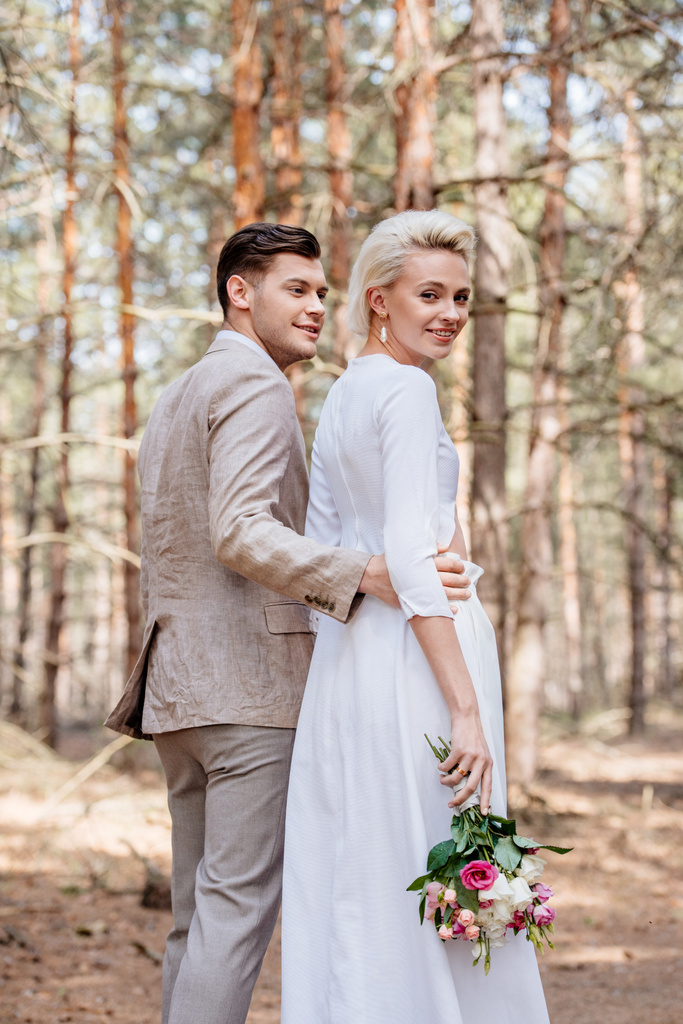 sposo sorridente in abbigliamento formale che abbraccia sposa nella foresta
 - Foto, immagini