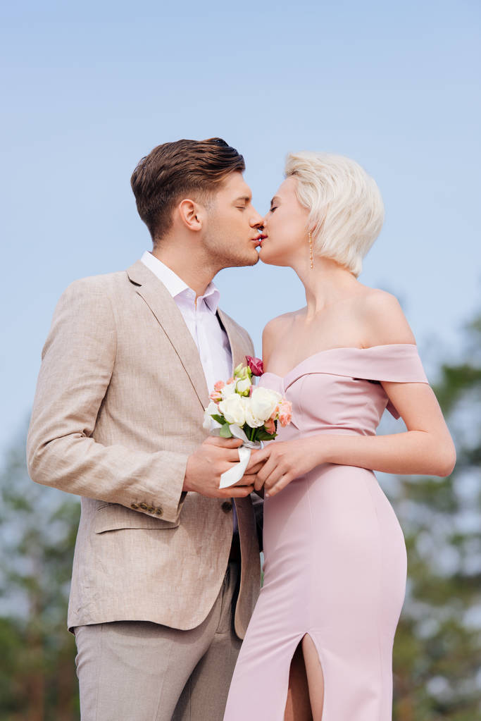 stilvolles Paar hält Brautstrauß und küsst sich mit geschlossenen Augen - Foto, Bild