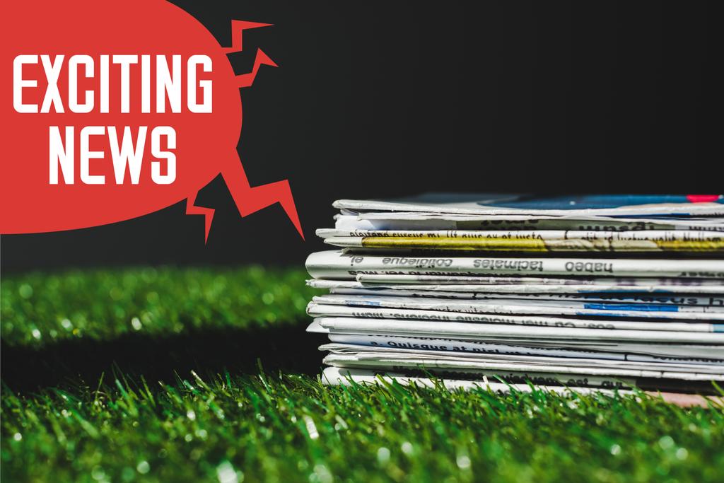 赤いスピーチバブルの近くの新鮮な緑の草の上に異なる印刷新聞のスタックと黒で隔離されたエキサイティングなニュースレタリング - 写真・画像