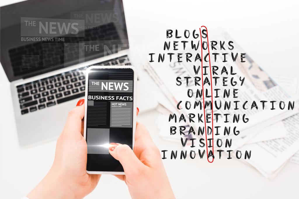 избирательный фокус женщины, держащей смартфон с иллюстрацией деловых новостей возле ноутбука, газет и кроссворда в социальных сетях
 - Фото, изображение