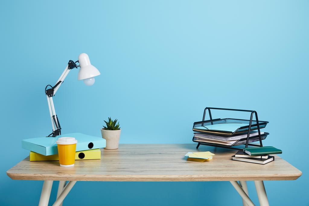 Arbeitsplatz mit Papieren, Sukkulente und Kaffee in Plastikbecher auf Holztisch auf blauem Hintergrund - Foto, Bild