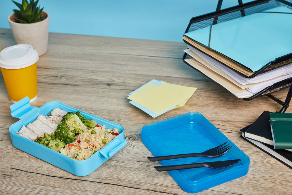 scatola da pranzo in plastica con cibo gustoso e sano sul posto di lavoro con carte su tavolo in legno su sfondo blu
 - Foto, immagini