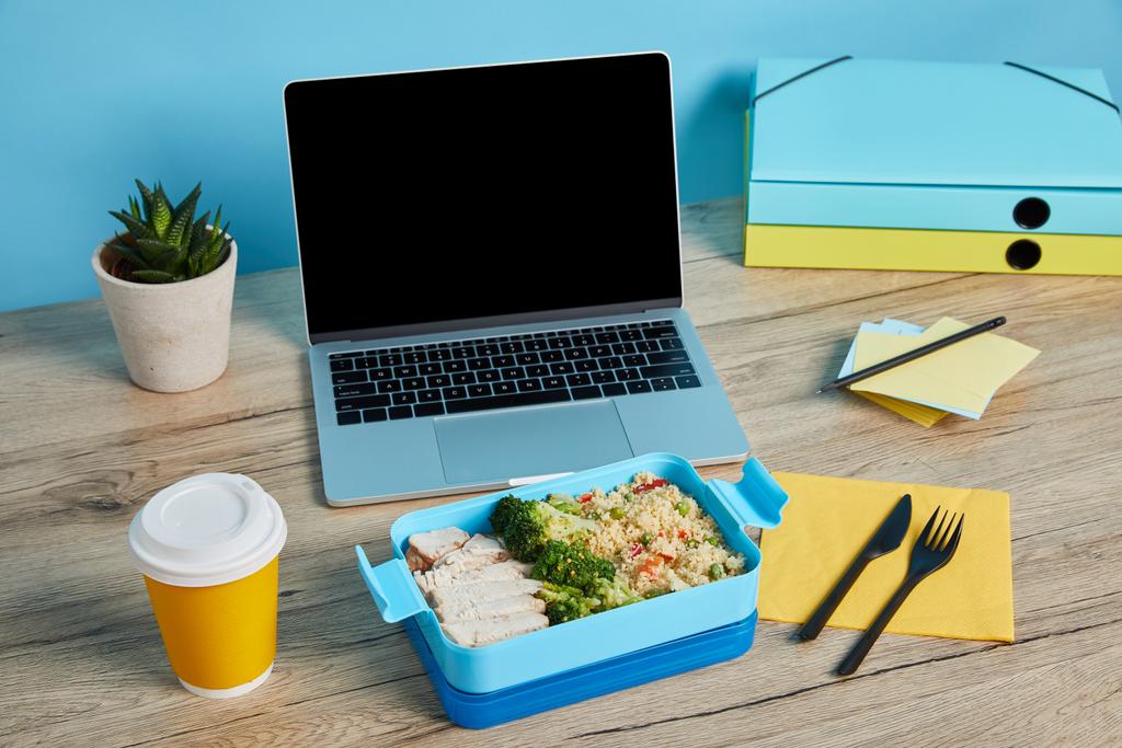 κουτί μεσημεριανό με ρύζι, κοτόπουλο και μπρόκολο στο χώρο εργασίας με φορητό υπολογιστή σε ξύλινο τραπέζι σε μπλε φόντο, ενδεικτικό συντακτική - Φωτογραφία, εικόνα