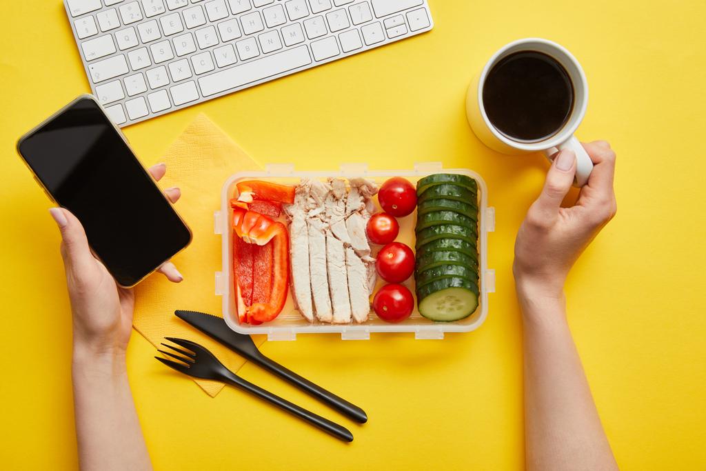 Vista cortada da mulher com delicioso almoço saudável segurando smartphone e xícara de café no fundo amarelo
  - Foto, Imagem