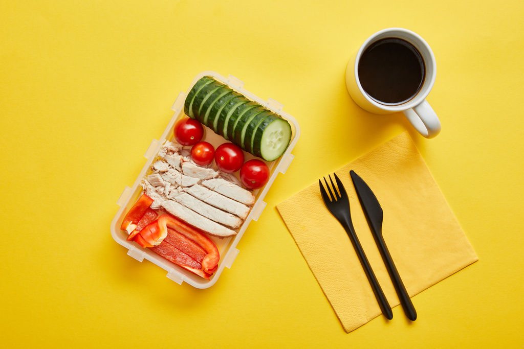 Top uitzicht van lunch box met kip en verse groenten en kopje koffie op gele achtergrond - Foto, afbeelding