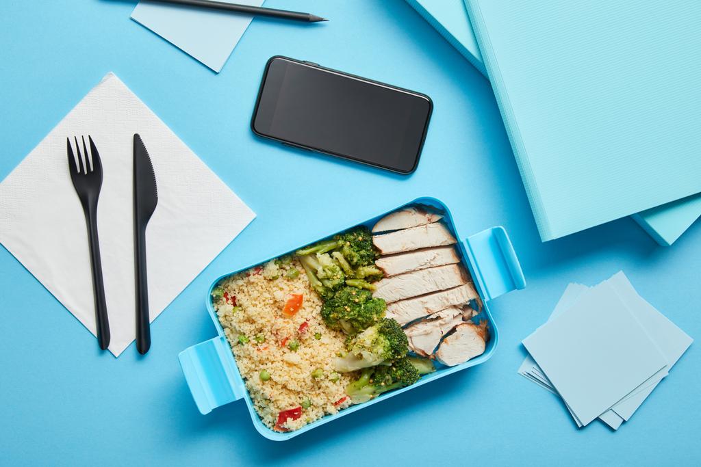 Κορυφαία θέα του κουτιού γεύμα με υγιεινό θρεπτικό φαγητό και smartphone στο χώρο εργασίας σε μπλε φόντο - Φωτογραφία, εικόνα