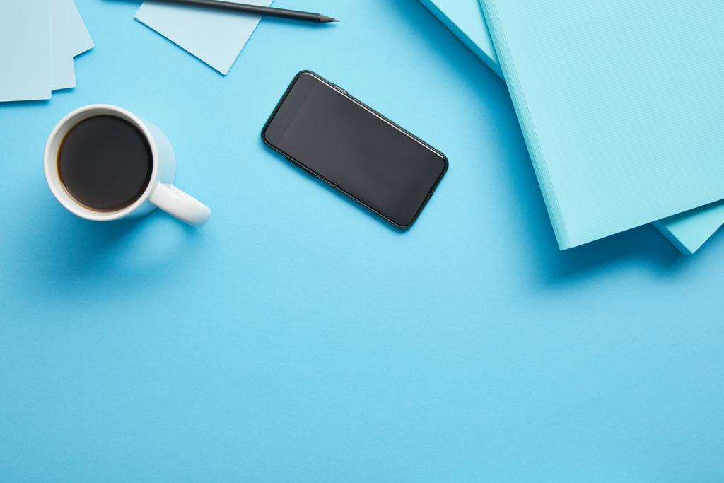 Bovenaanzicht van smartphone met blanco scherm en kopje koffiezet op de werkplek op een blauwe achtergrond - Foto, afbeelding
